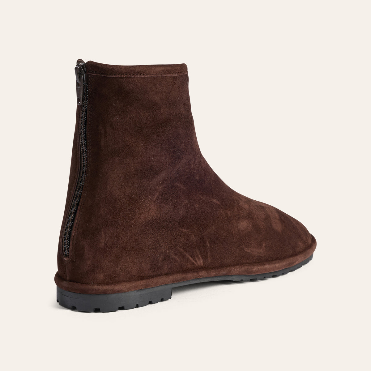 A dark brown suede ankle boot with a side zipper and a black lug sole, shown from a three-quarters angle against a light cream background.
