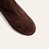 A close-up of a dark brown suede boot showing the toe and side profile against a light cream background.