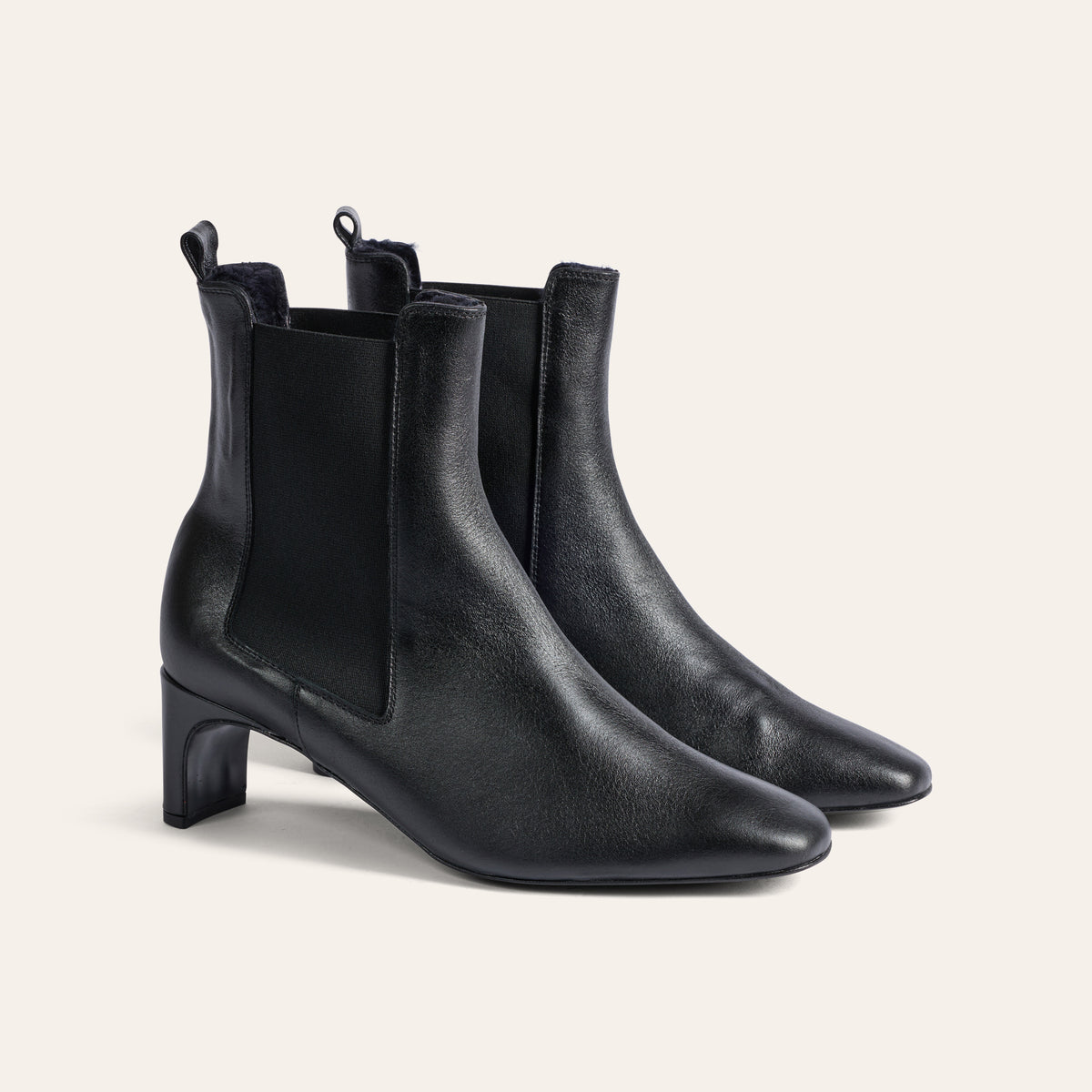 A pair of black leather ankle boots with pointed toes, mid-height block heels, and elastic side panels, shown on a light beige background.