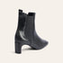 A black leather ankle boot with a block heel and elastic side gore, shown from a three-quarters rear angle on a light cream background.