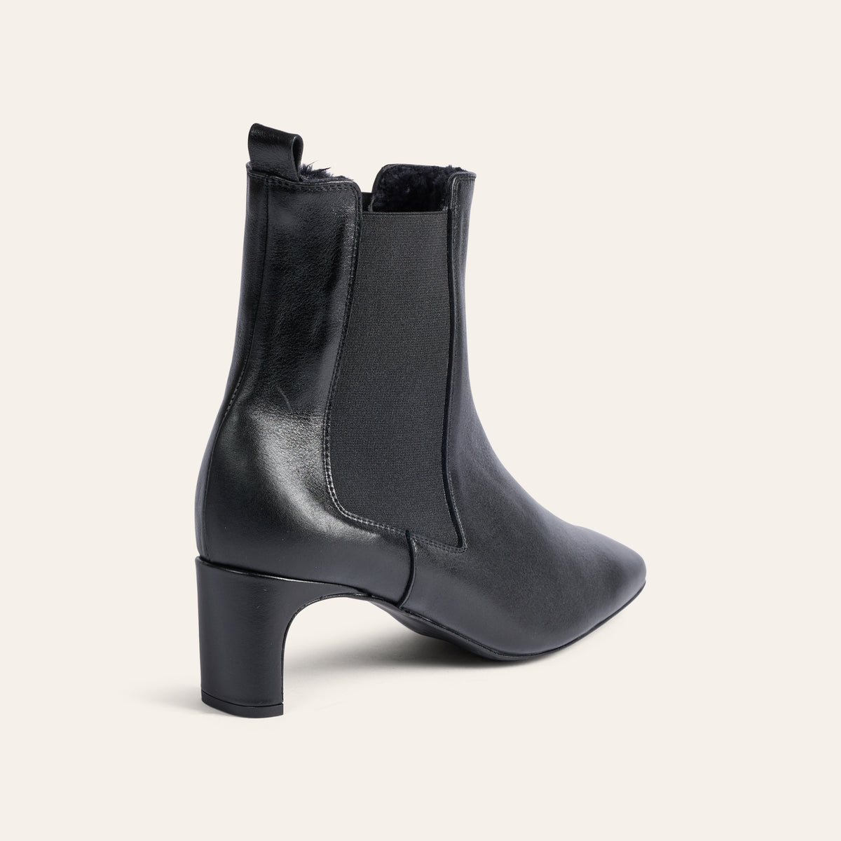 A black leather ankle boot with a block heel and elastic side gore, shown from a three-quarters rear angle on a light cream background.