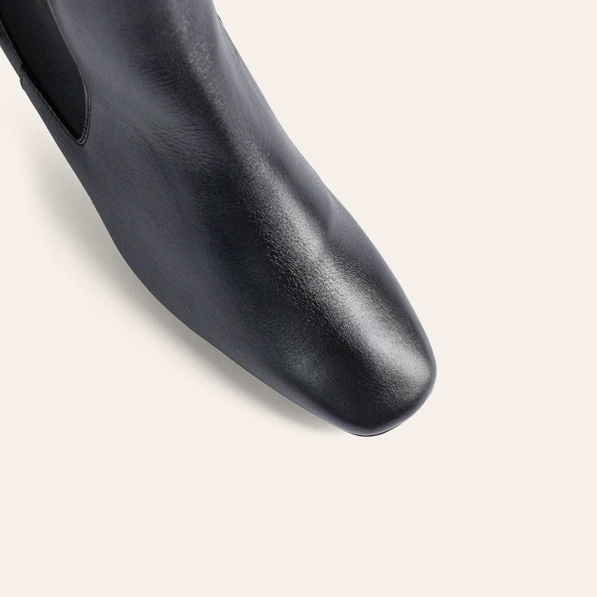 Close-up of a black leather boot with a square toe and a side cutout, shown on a light beige background.