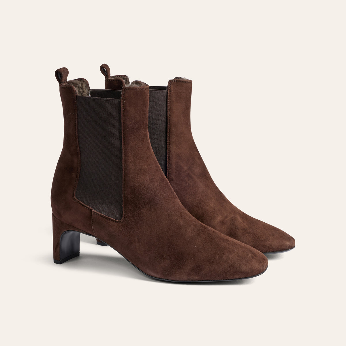 A pair of dark brown suede Chelsea-style ankle boots with black elastic side panels and low block heels, shown on a light cream background.