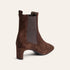 A brown suede Chelsea-style ankle boot with a block heel and a dark brown elastic side panel, shown from a three-quarter rear angle on a light cream background.