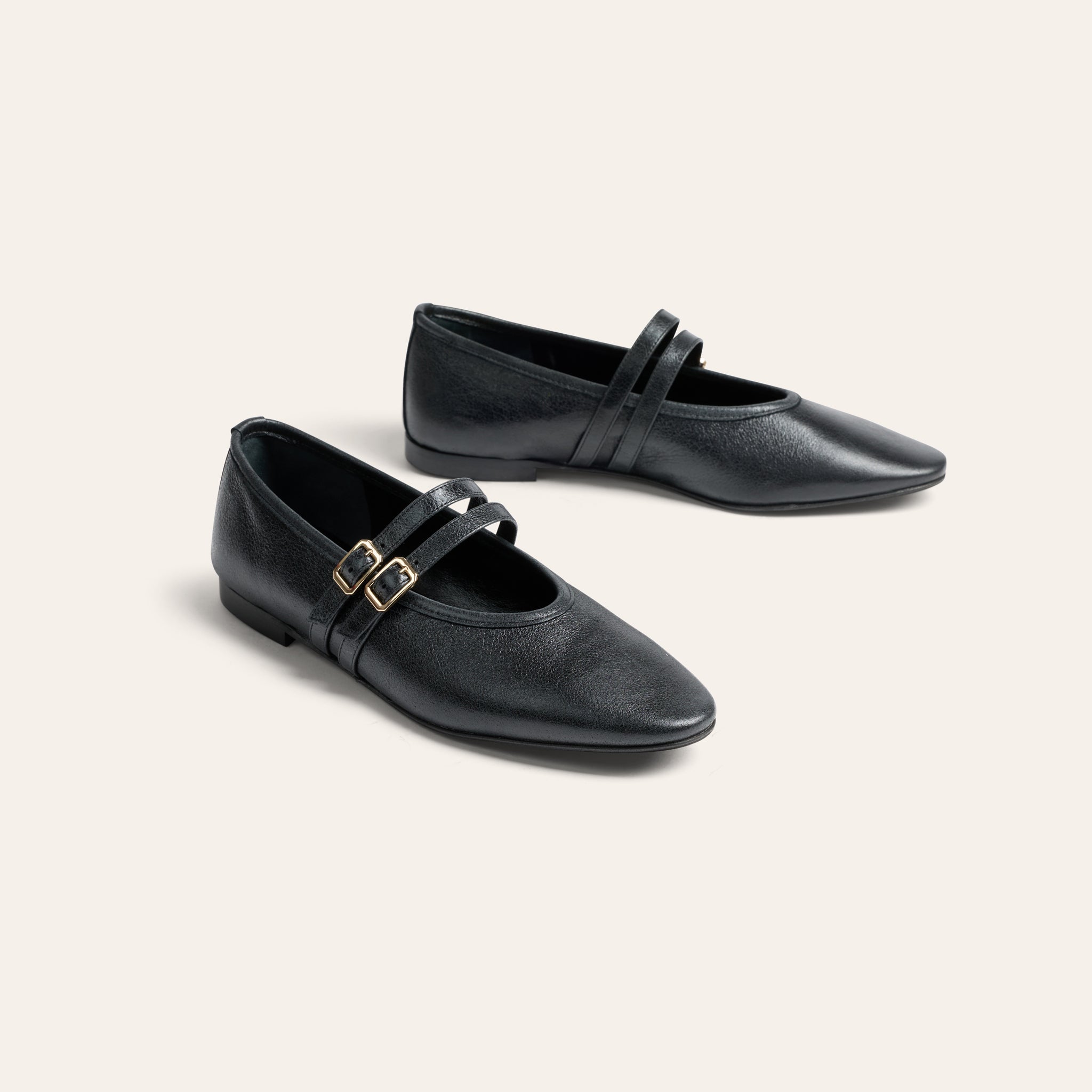 A pair of black leather Mary Jane flats with pointed toes and double straps accented by gold-tone buckles, displayed on a light beige background.
