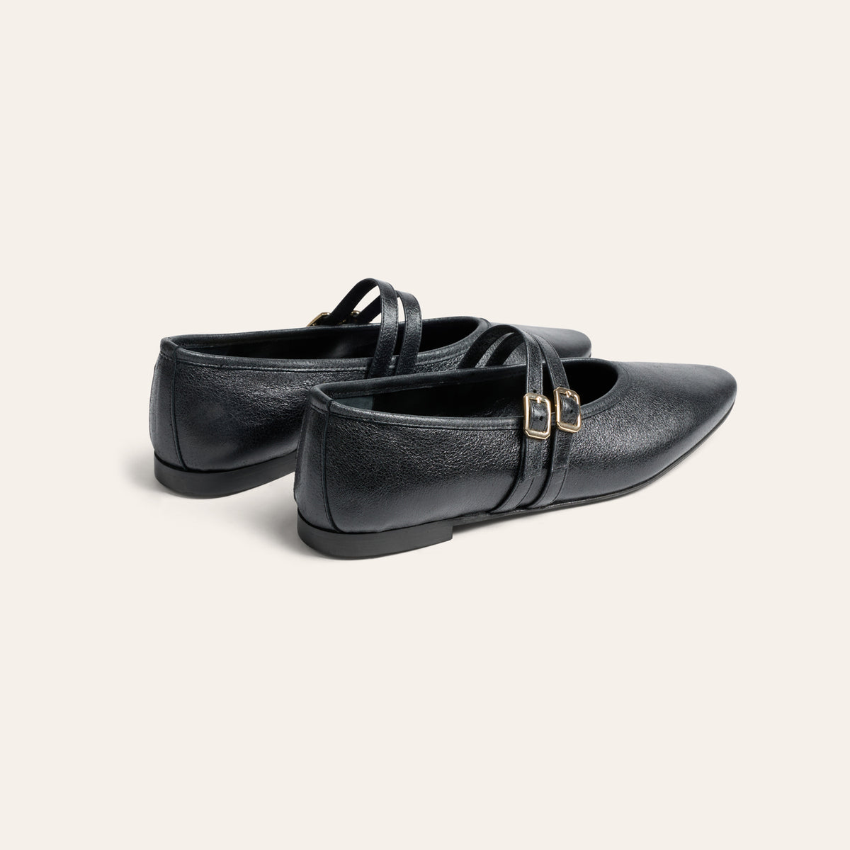 A pair of black leather Mary Jane flats with double straps and small gold-tone buckle details, shown from a three-quarters angle on a light cream background.