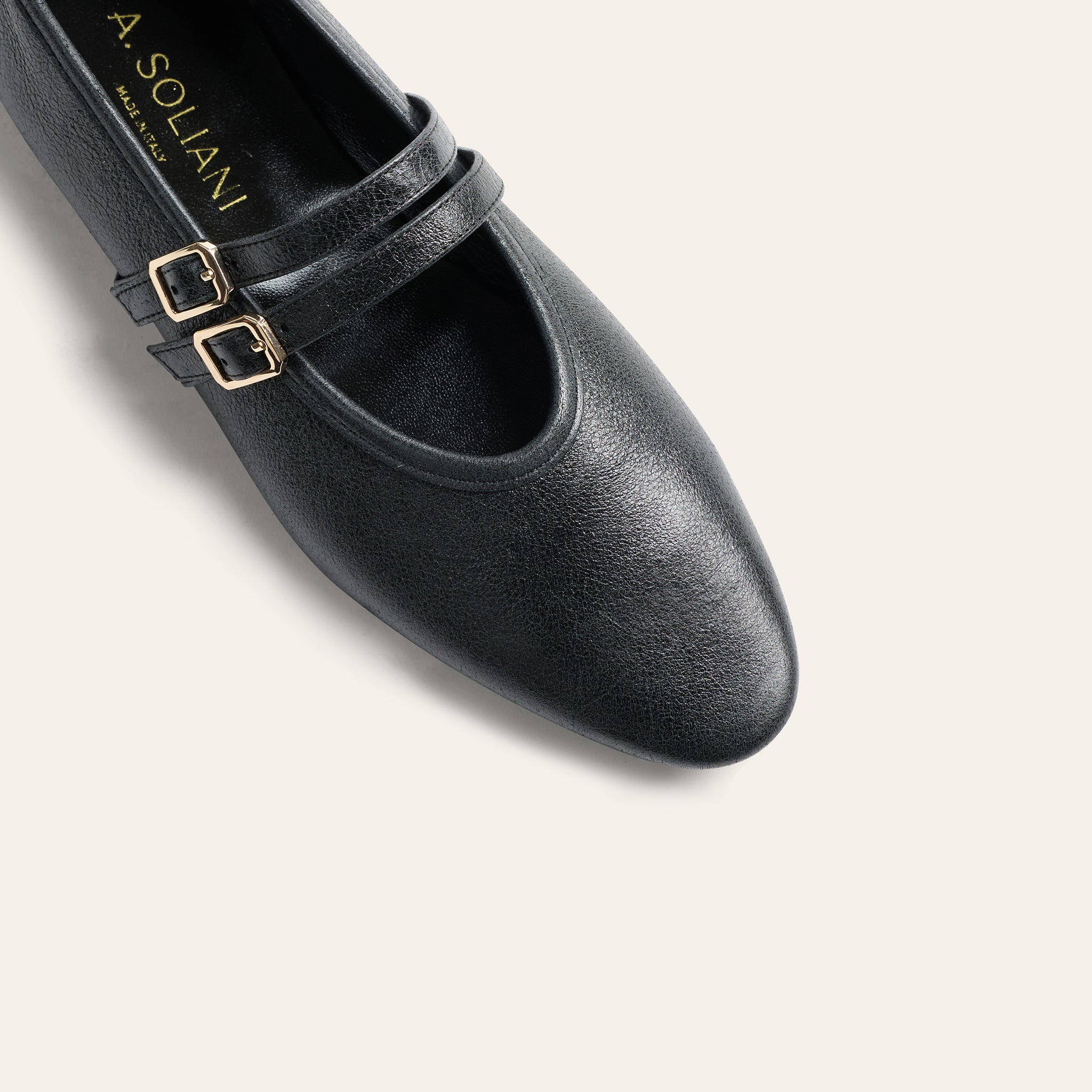 A close-up studio shot of a black leather Mary Jane shoe by A. Soliani, featuring a double strap design with gold-tone buckle details, set against a solid off-white background.
