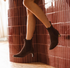 A person wearing dark brown suede Chelsea-style ankle boots with mid-height block heels, posed against a glossy reddish-brown tiled wall.