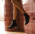 A person wearing black ankle boots with low block heels, photographed indoors against a glossy pink-brown tiled wall.