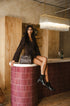 A woman sits on a cylindrical bar counter in a rustic indoor setting, wearing a dark brown long-sleeve top and black ankle boots, with a structured dark brown handbag resting beside her.