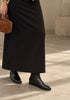 A person wearing a long black midi skirt, black leather ankle boots, and holding a brown leather handbag in a minimalist indoor setting with concrete floors.