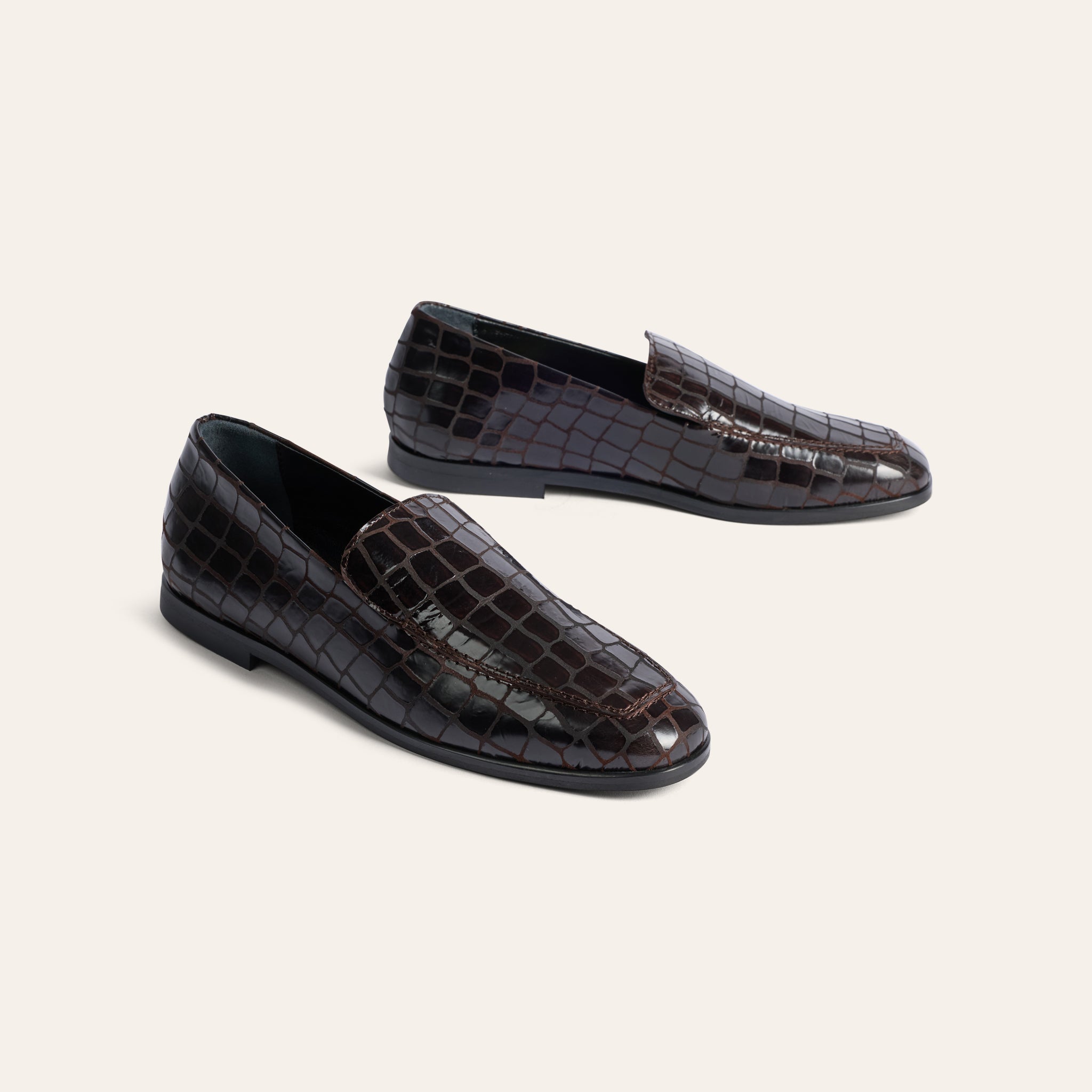 A pair of dark brown crocodile-embossed leather loafers on a light beige background.