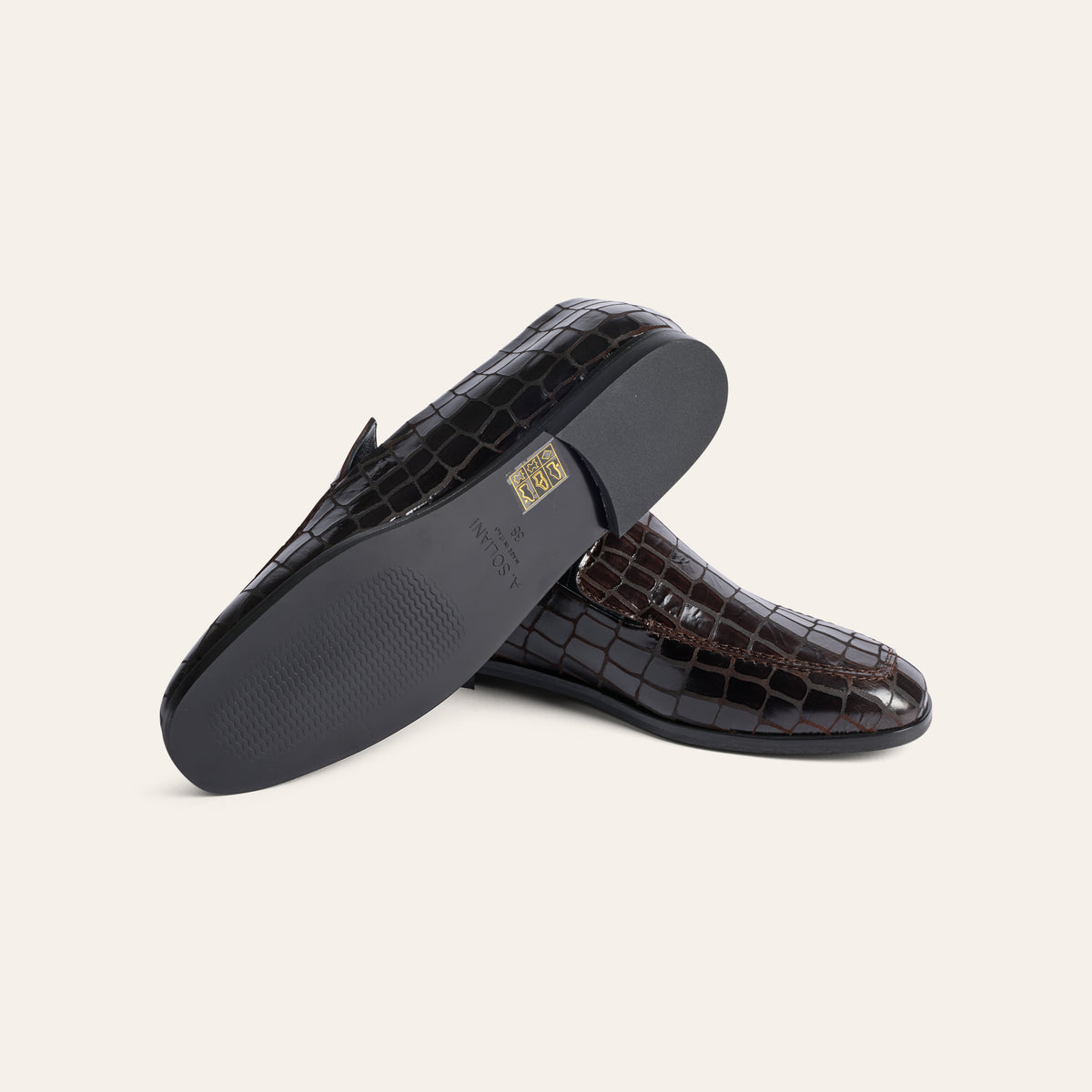 A pair of dark brown crocodile-embossed leather loafers, with one shoe turned to show the black outsole and a small gold brand plaque, set against a solid off-white background.