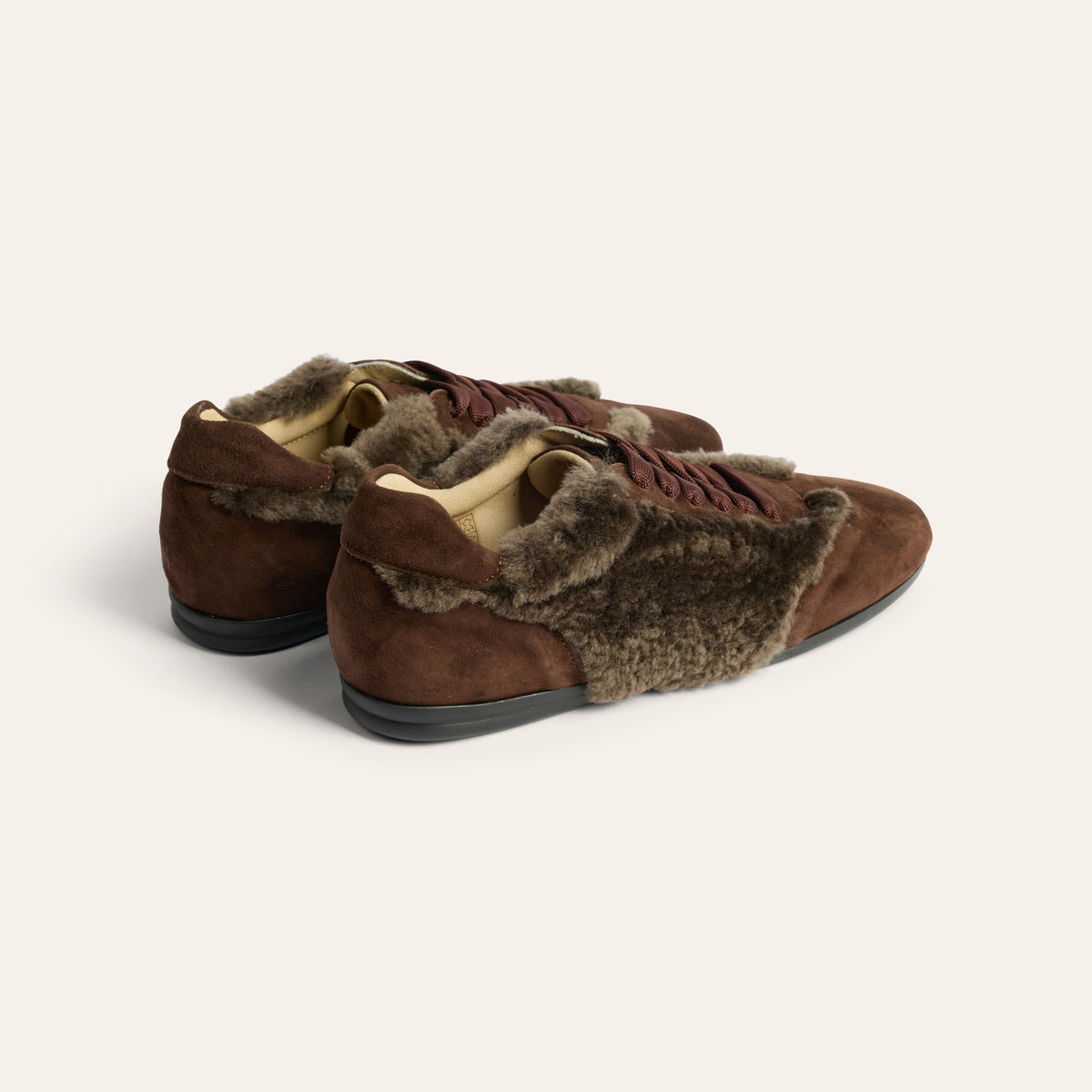 A pair of brown suede and shearling slip-on shoes with dark brown laces, shown from a three-quarters angle on a light beige background.