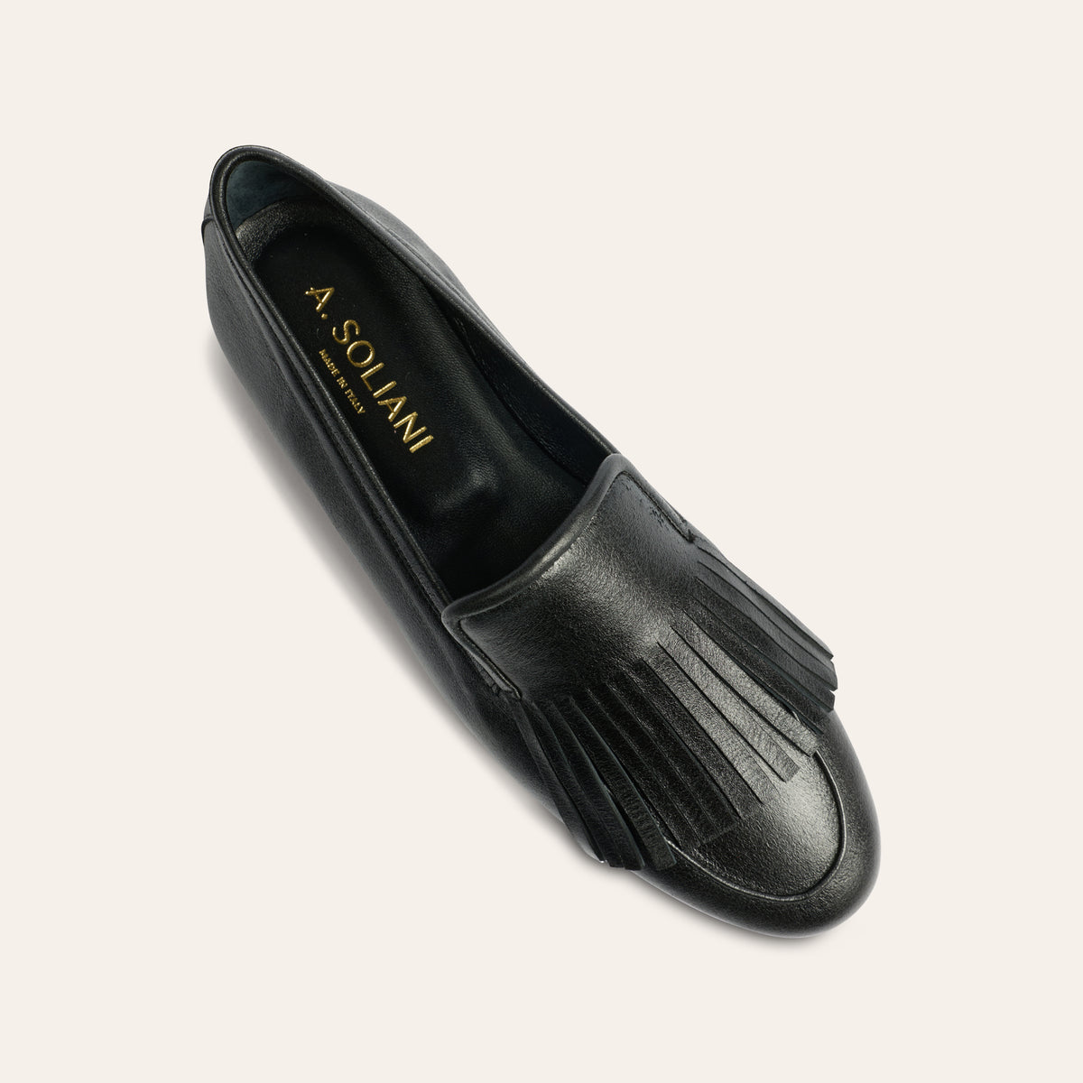 A single black leather loafer with fringe detailing on the toe, shown from a top-down angle on a light beige background. The brand name 'A.