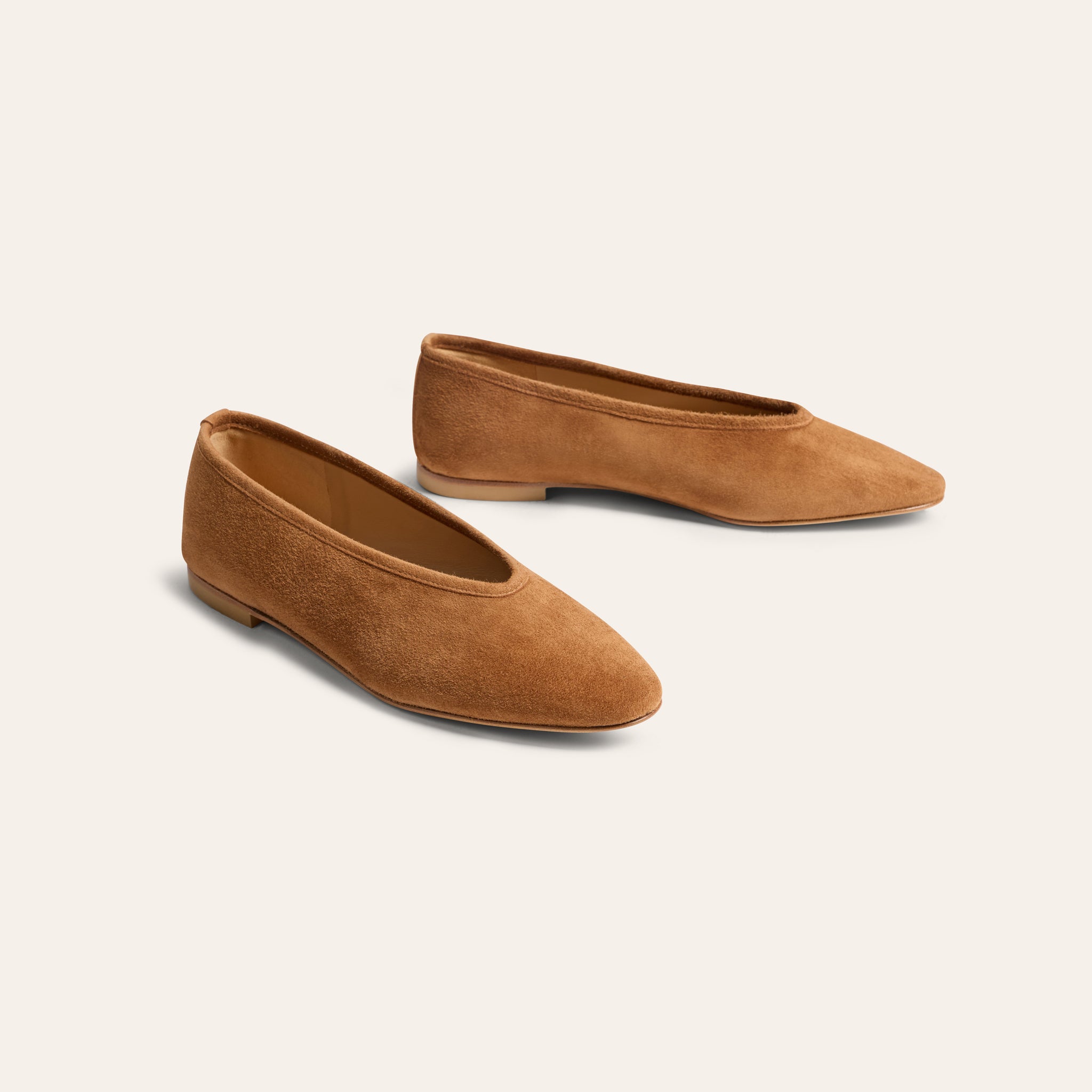 A pair of tan suede flat shoes with pointed toes, displayed on a clean, solid off-white background.