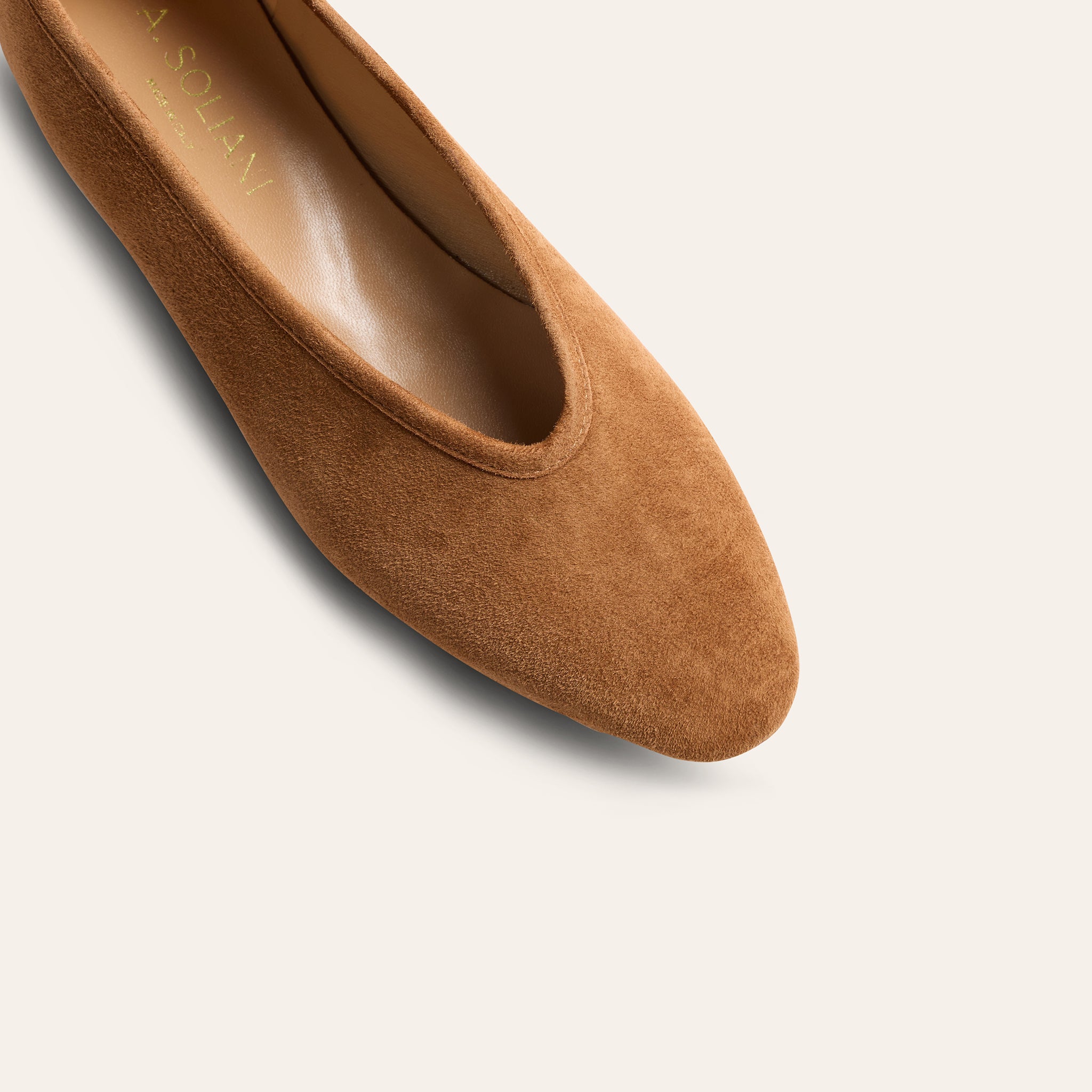 A close-up shot of a tan suede flat shoe, showing the pointed toe and the interior insole with the brand name printed in gold.