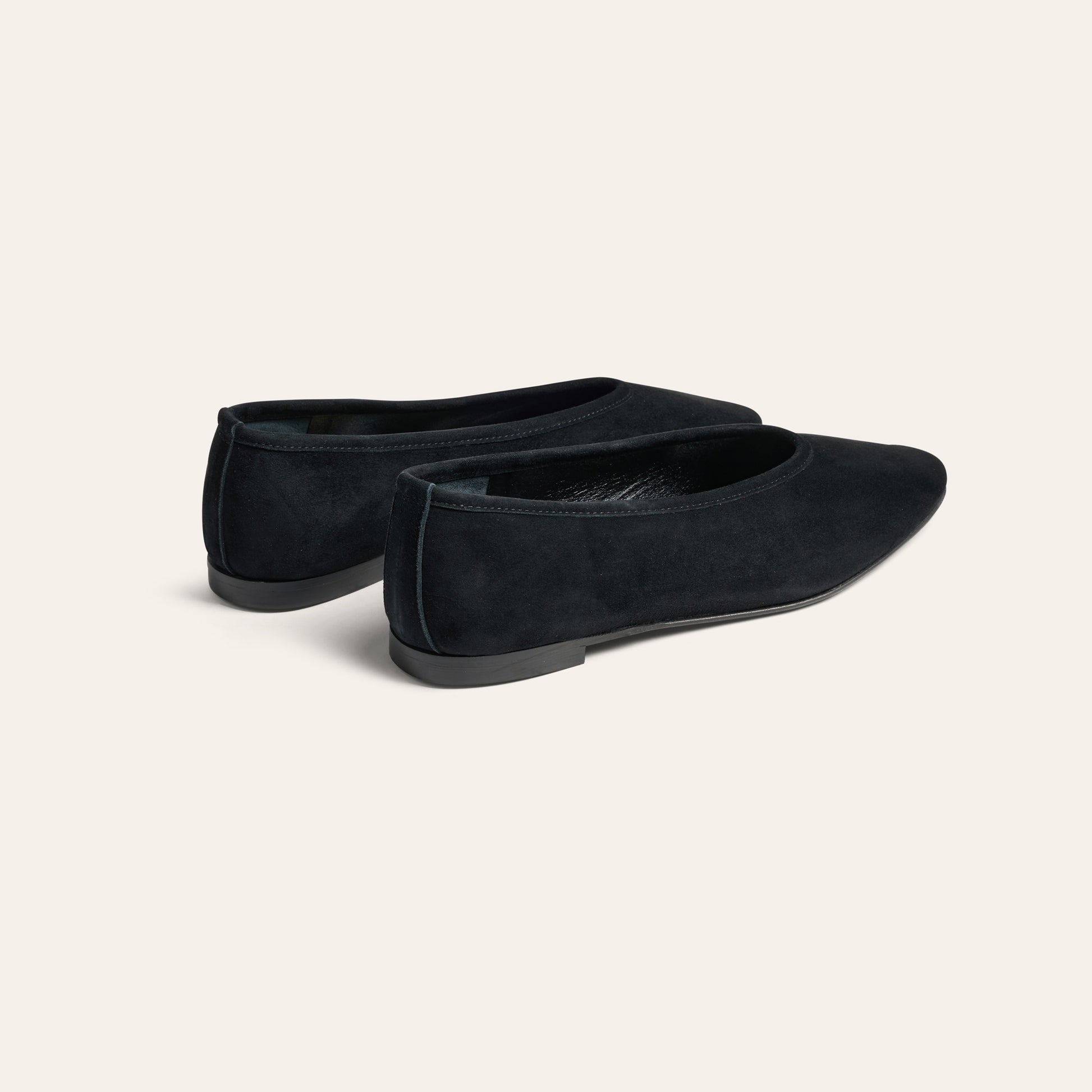 A pair of black suede flat shoes with pointed toes, shown from a three-quarters angle on a light cream background.