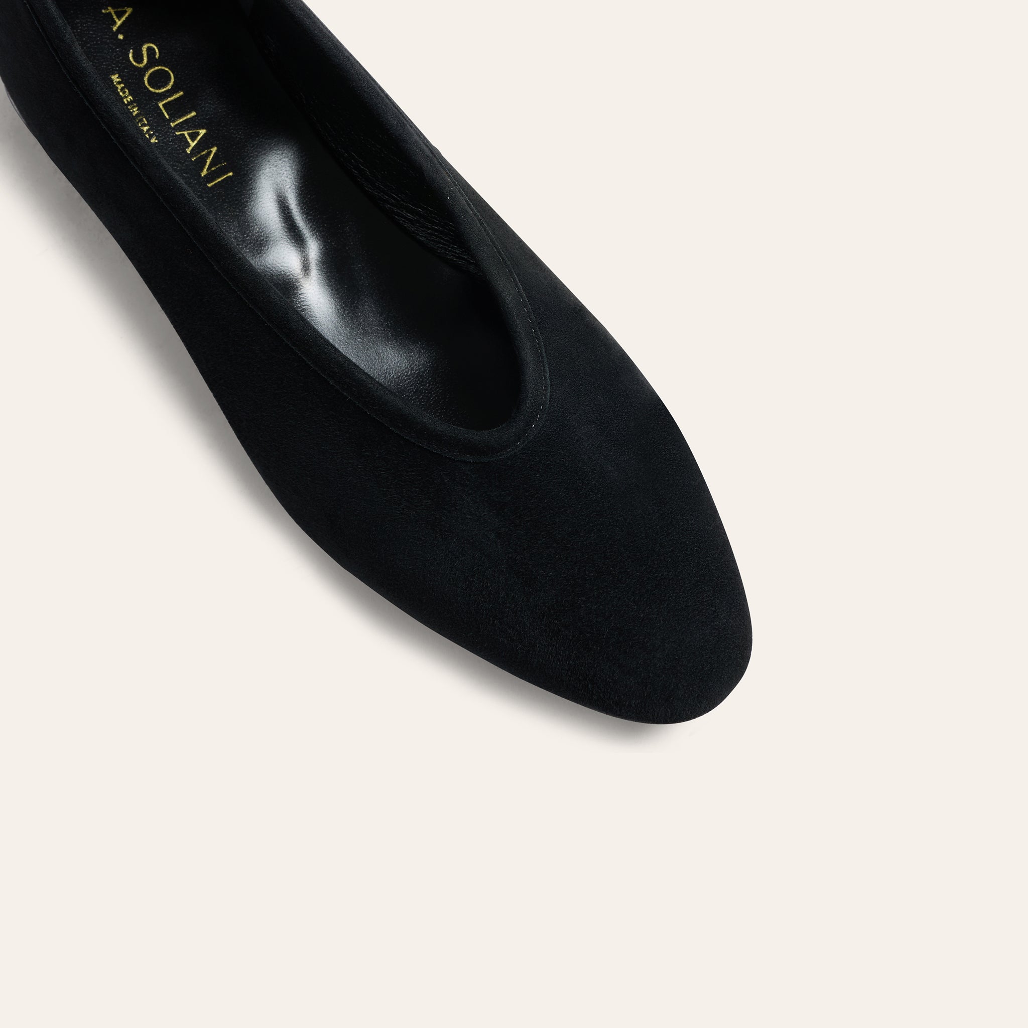 A close-up studio shot of a black pointed-toe flat shoe by A. Soliani, showing the black suede-like exterior and the gold brand name printed inside the heel area, set against a solid off-white background.