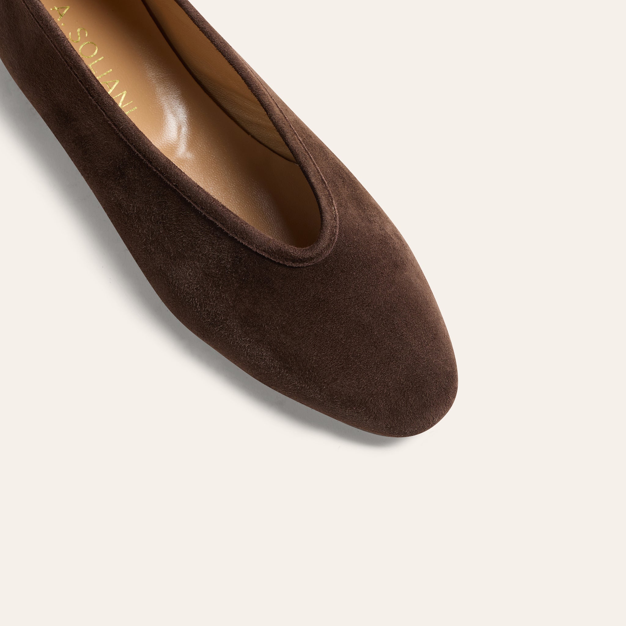 A close-up of a dark brown suede flat shoe, showing the textured suede upper, rounded toe, and tan insole with gold lettering, set against a solid off-white background.