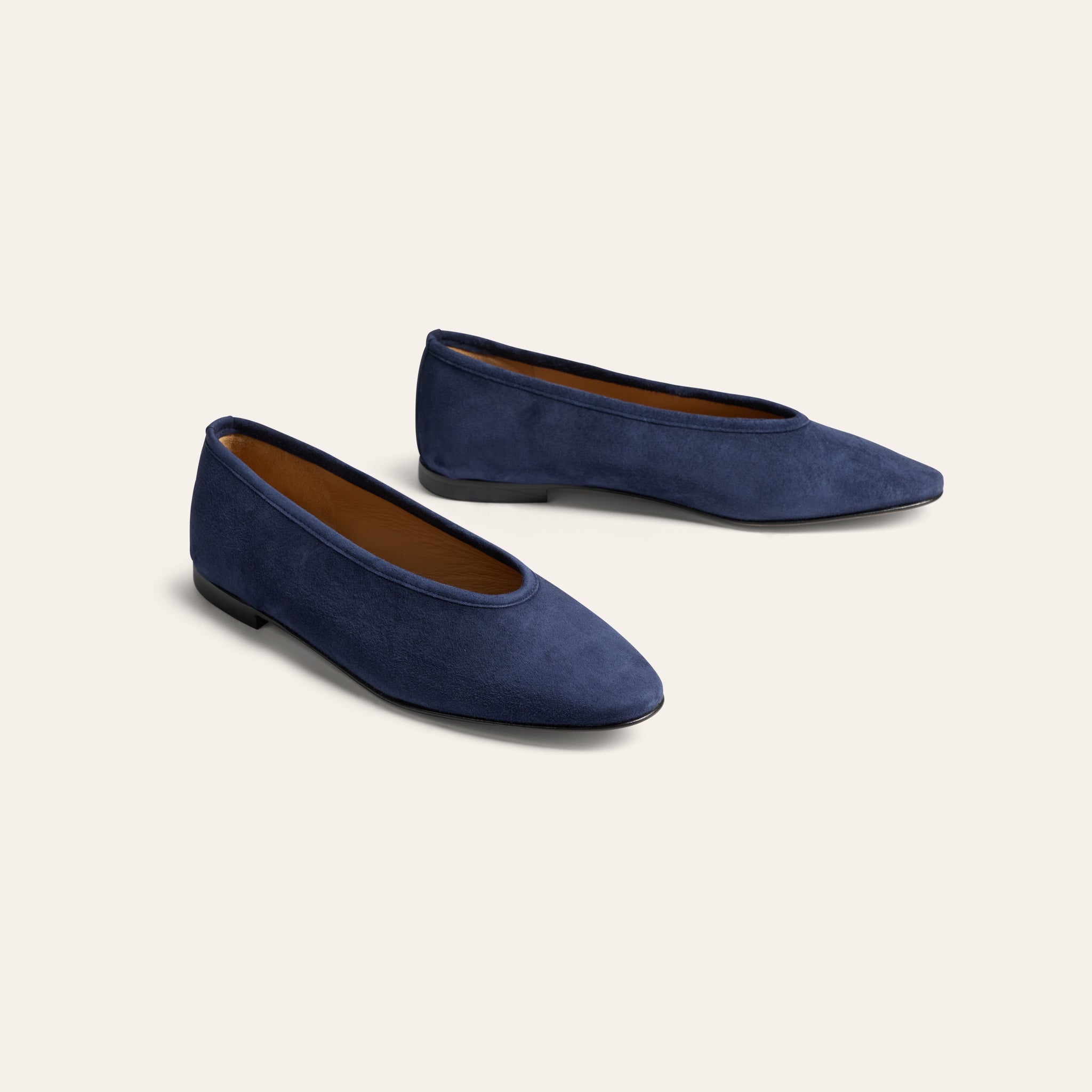 A pair of navy blue suede pointed-toe flats with a tan interior lining, displayed on a clean, light beige background.
