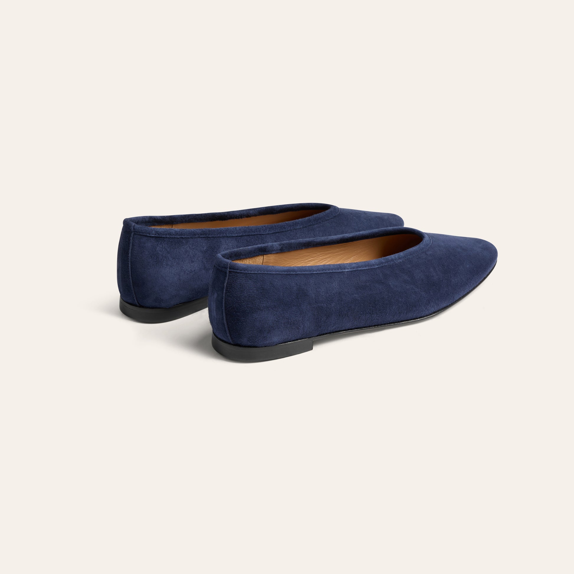 A pair of navy blue suede flat shoes with black soles, shown from a three-quarters angle on a light beige background.