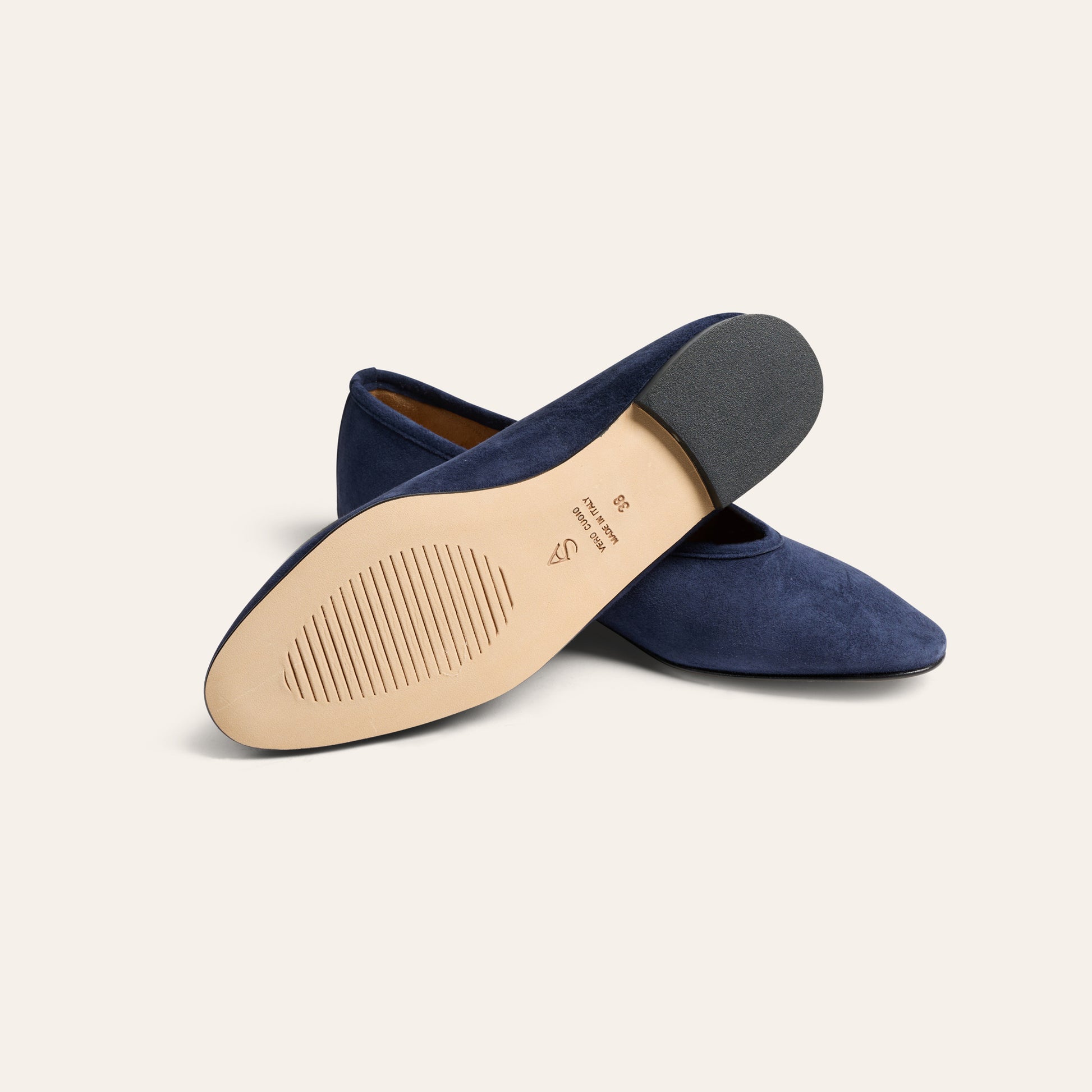 A pair of navy blue suede pointed-toe flats, with one shoe turned over to show the beige insole and dark outsole, set against a solid off-white background.
