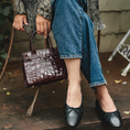 Load image into Gallery viewer, Person wearing a patterned coat, blue jeans, and black shoes, holding a brown handbag.
