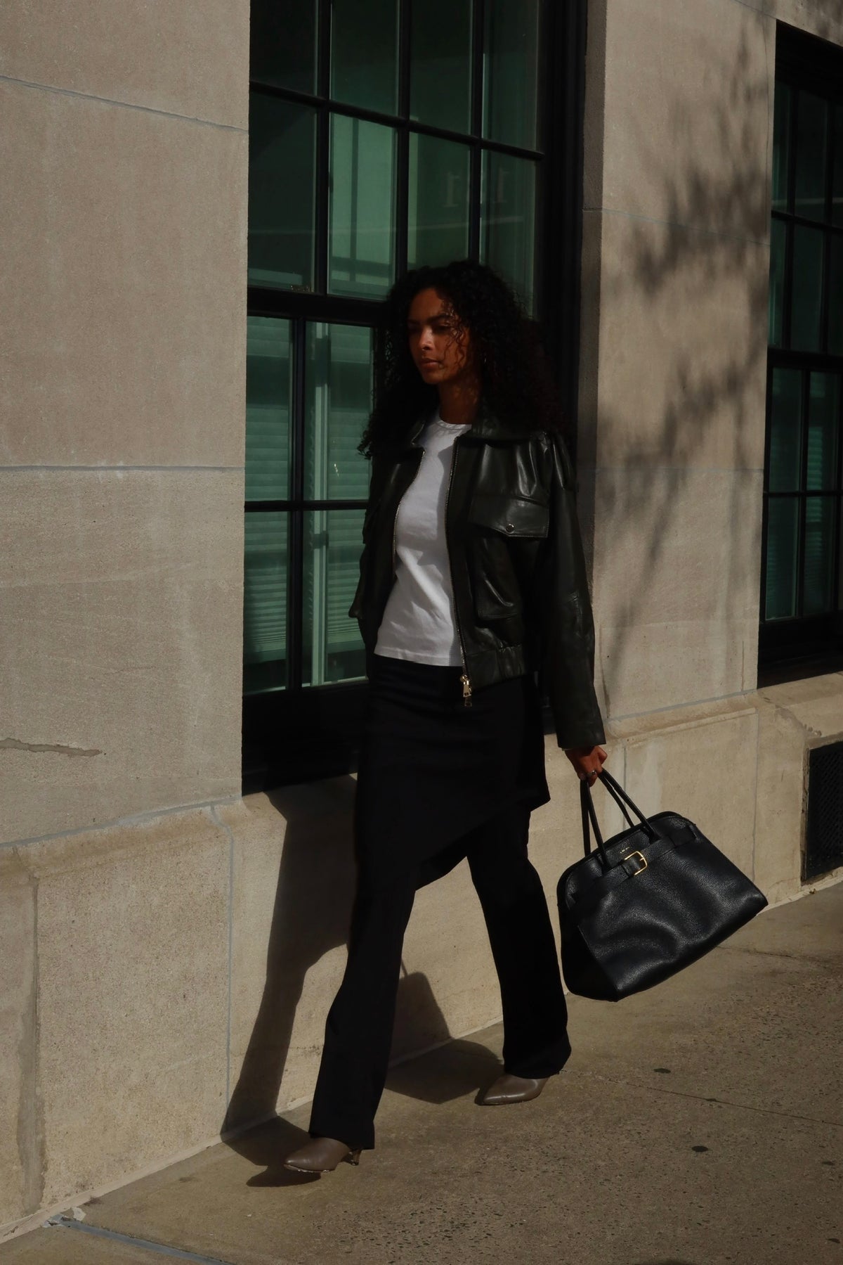 A woman walking outdoors next to a stone building, wearing a green leather jacket, white top, black pants, and carrying a black leather handbag.