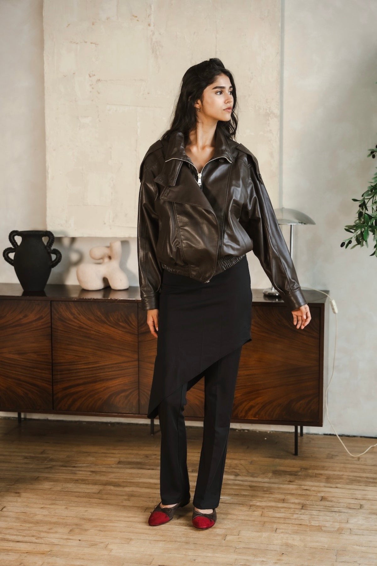 A woman stands in a room wearing a dark brown leather jacket, a black asymmetrical skirt, black trousers, and red shoes. She is posed in front of a wooden cabinet and textured wall.